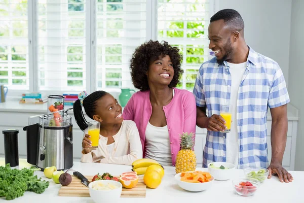 Sonrientes padres e hija tomando un vaso de jugo de naranja en la cocina en casa — Foto de Stock