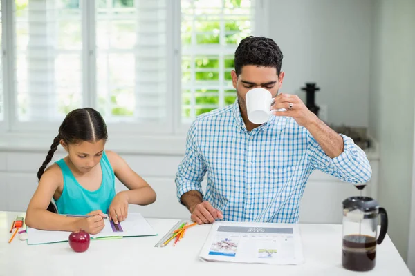 Hija haciendo su tarea y padre tomando una taza de té negro mientras lee el periódico — Foto de Stock