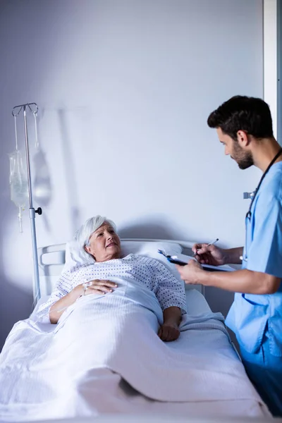 Male doctor discussing medical report with female senior patient on bed — Stock Photo, Image