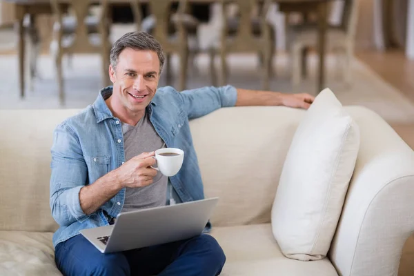 Портрет улыбающегося мужчины, сидящего на диване с ноутбуком, пьющего кофе — стоковое фото