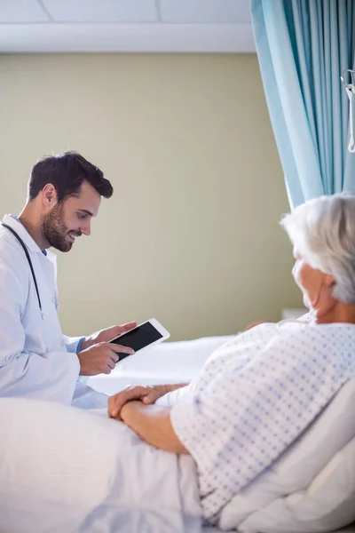 Врач-мужчина обсуждает медицинский отчет за цифровым планшетом со старшей пациенткой в палате — стоковое фото