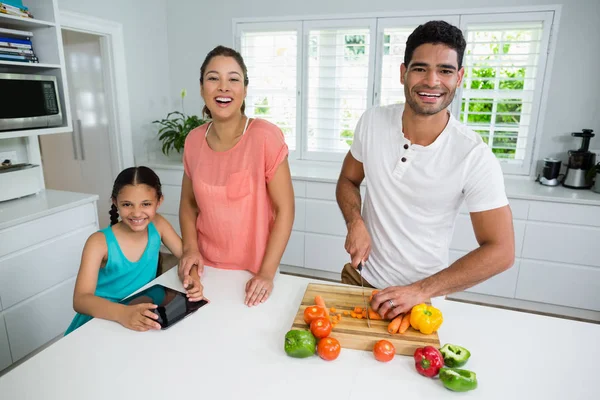 Πορτρέτο της κόρης και την μητέρα χρησιμοποιώντας ψηφιακό tablet, ενώ ο πατέρας κόψιμο λαχανικών στην κουζίνα — Φωτογραφία Αρχείου