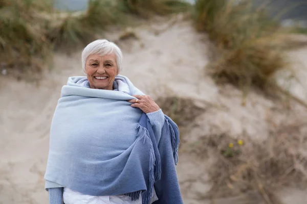 Femme âgée souriante enveloppée dans un châle à la plage — Photo