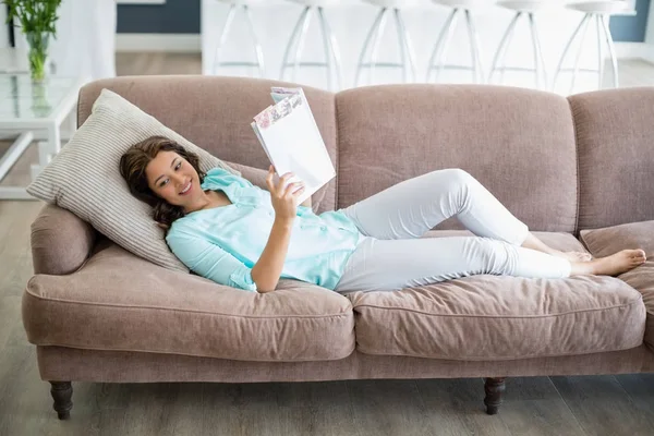 Mujer sonriente leyendo revista mientras está acostado en el sofá en la sala de estar —  Fotos de Stock