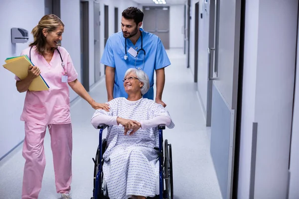 Médico masculino y femenino interactuando con paciente senior femenino en silla de ruedas en corredor — Foto de Stock