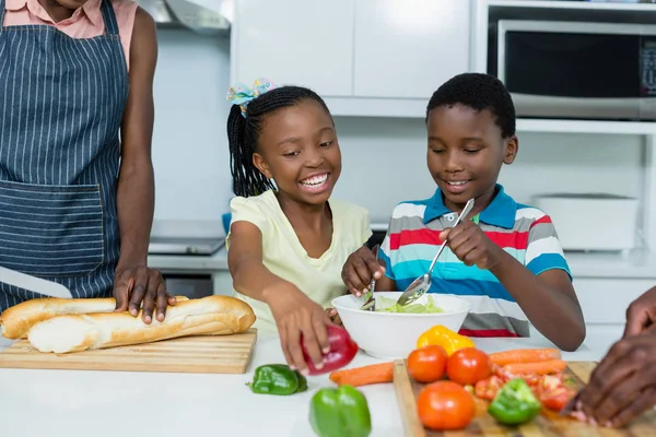 Kids preparing salad while mother cutting bread loaf in kitchen — Stock Photo, Image