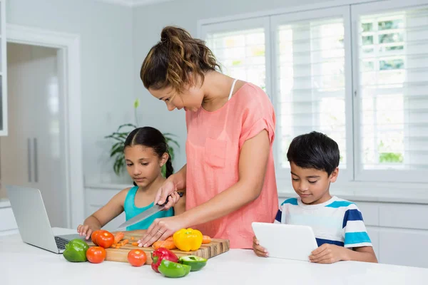 Niños usando tableta digital y computadora portátil mientras la madre corta verduras en la cocina — Foto de Stock