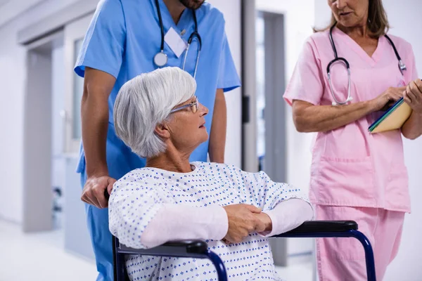 Médico masculino y femenino interactuando con paciente senior femenino en silla de ruedas en corredor —  Fotos de Stock