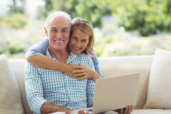 Porträt einer lächelnden Seniorin, die einen Mann im Wohnzimmer umarmt, während sie einen Laptop benutzt — Stockfoto