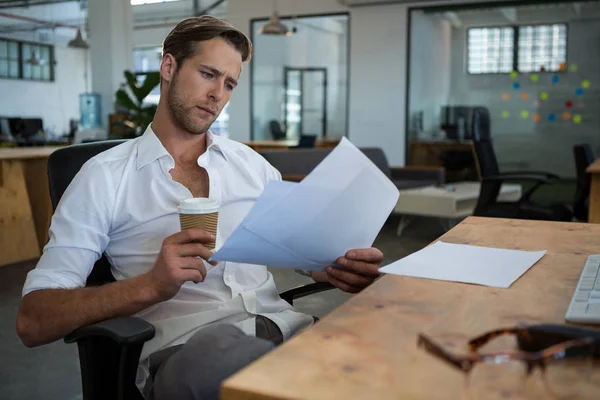 Biznesmen, trzymając kubek kawy jednorazowe podczas czytania dokumentu o biurko — Zdjęcie stockowe