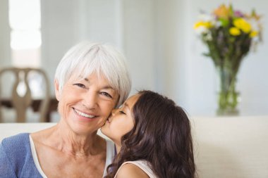 Granddaughter kissing grandmother on cheek in living room clipart