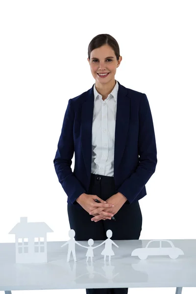 Усміхнена бізнес-леді, що стоїть з папером, вирізаним з сім'ї, будинку та автомобіля на столі — стокове фото