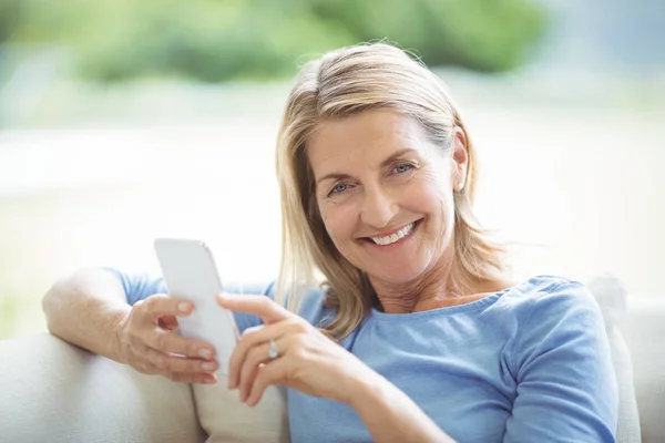 Uśmiechnięta kobieta senior przy użyciu telefonu komórkowego w salonie — Zdjęcie stockowe