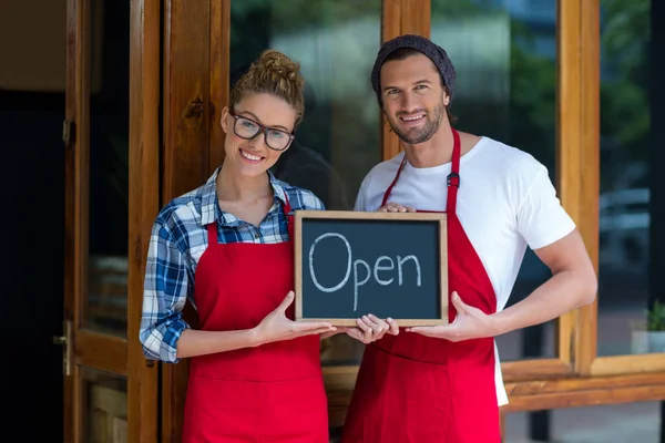 Smiling waitress and waiter standing with open sign board outside cafe — Stock Photo, Image