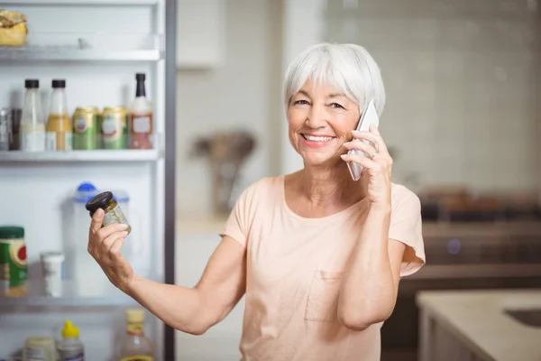 Senior woman looking at jar while talking on mobile phone in kitchen — Stock Photo, Image