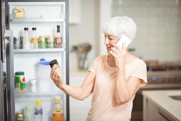 Senior kvinna som tittar på burken medan du talar i mobiltelefon i köket — Stockfoto