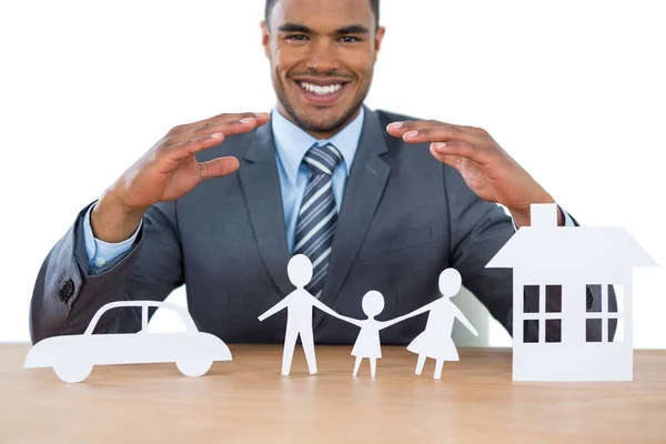 Smiling businessman protecting paper cut out family, house and car with hands — Stock Photo, Image
