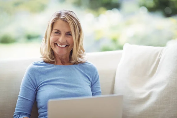 Donna anziana sorridente seduta sul divano con laptop in soggiorno — Foto Stock