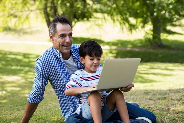 Boy sitting on his fathers lap and using laptop in park — Stock Photo, Image