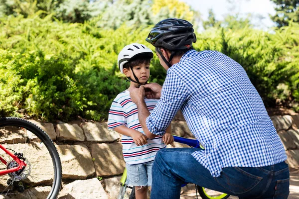 Father assisting son in wearing bicycle helmet in park — Stock Photo, Image