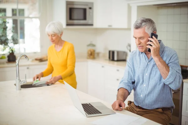 Senior man praten op mobiele telefoon terwijl vrouw wassen plaat in keuken — Stockfoto