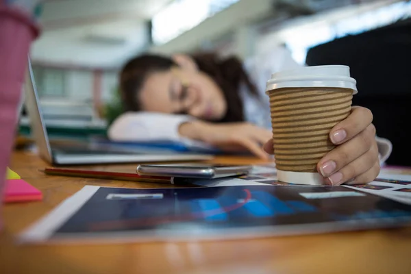 Tek kullanımlık bardak masanın üzerinde uyurken tutarak yorgun kadın grafik tasarımcı — Stok fotoğraf