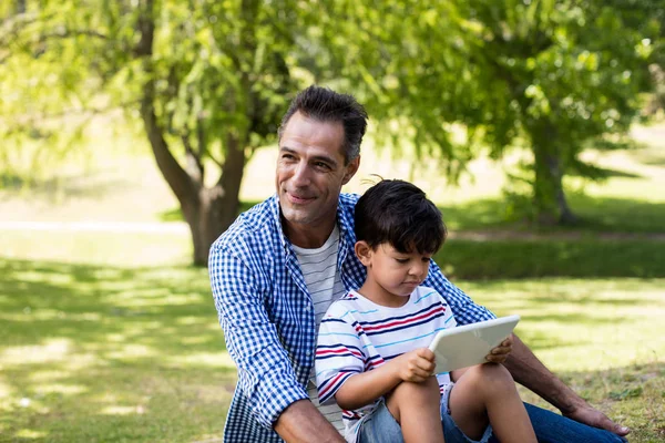 Garçon assis sur les genoux de son père et utilisant une tablette numérique dans le parc — Photo