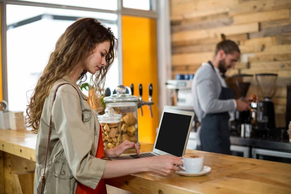 Γυναίκα στέκεται στο μετρητή και να χρησιμοποιούν φορητό υπολογιστή ενώ έχοντας καφέ — Φωτογραφία Αρχείου