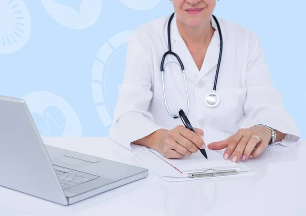 Женщина-врач сидит за столом с ноутбуком и пишет на планшете — стоковое фото