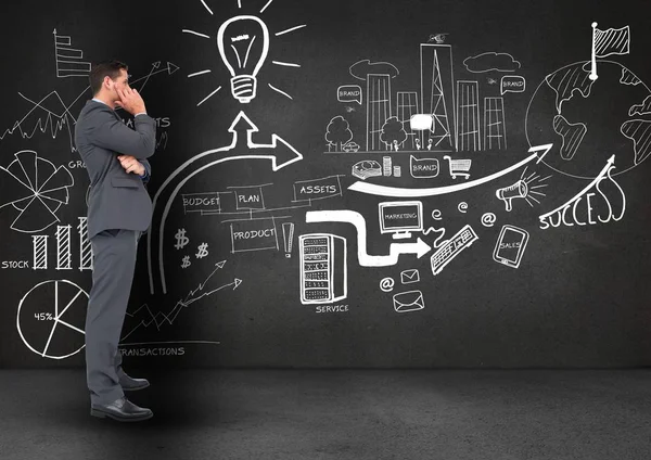 Thoughtful businessman standing against business symbols on blackboard — Stock Photo, Image