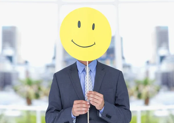 Uomo d'affari che tiene felice faccina sorridente davanti alla sua faccia contro ufficio in background — Foto Stock