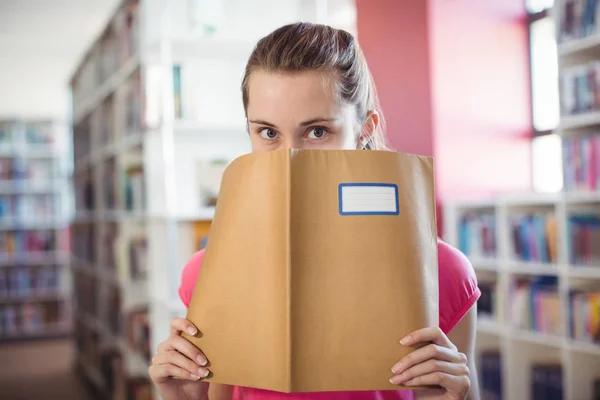 Schoolgirl hiding her face with book in library — Stock Photo, Image