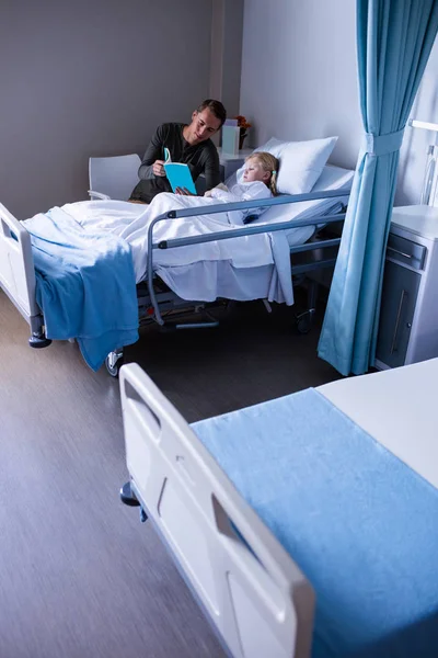 Girl on hospital bed reading book with father — Stock Photo, Image