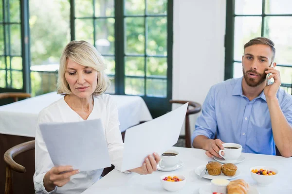 Female executive looking at document while colleague talking on mobile phone — Stock Photo, Image