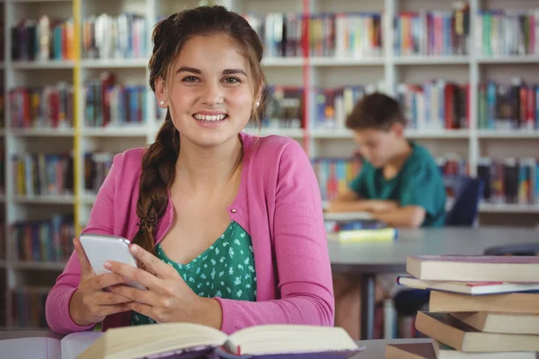 Smiling schoolgirl using mobile phone in library at school — Stock Photo, Image