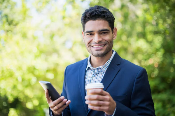 Empresario sosteniendo taza de café desechable y teléfono móvil — Foto de Stock