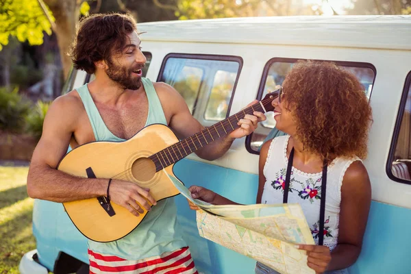 Mies soittaa kitaraa lähellä campervan ja nainen tilalla kartta hänen vieressään — kuvapankkivalokuva