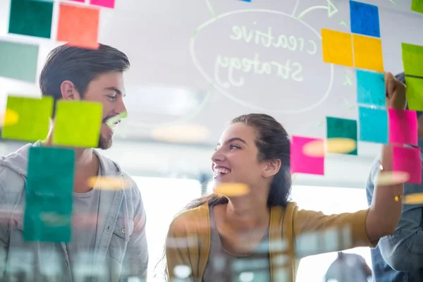 Lachende leidinggevenden interactie met elkaar tijdens het schrijven op plaknotities op glazen wand — Stockfoto