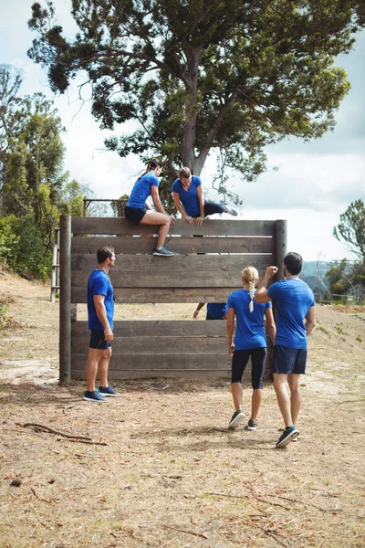 Entraîneur féminin aidant femme en forme à grimper au-dessus du mur en bois pendant le parcours d'obstacles — Photo