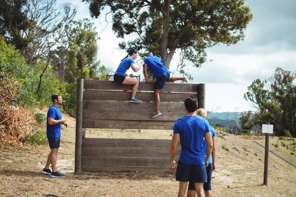 Entraîneur féminin aidant l'homme en forme à grimper au-dessus du mur en bois pendant le parcours d'obstacles — Photo