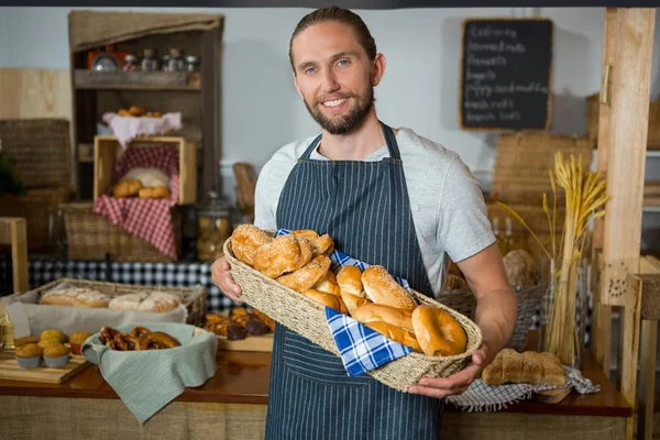 Smiling male staff holding wicker basket of various breads at counter — Stock Photo, Image