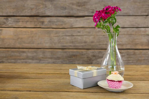 Scatola regalo e vaso di fiori con cupcake in piatto su tavola di legno — Foto Stock