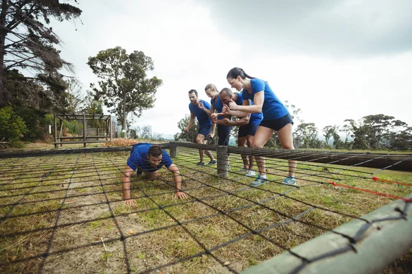 Hombre en forma arrastrándose debajo de la red durante la carrera de obstáculos, mientras que la gente en forma animando —  Fotos de Stock