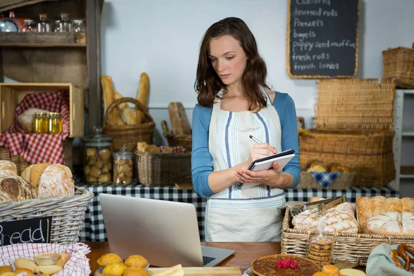 Personalen tittar på laptop medan du skriver i dagboken i bageri counter — Stockfoto