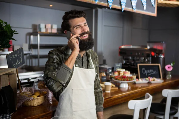 Smiling male staff talking on mobile phone at counter in coffee shop — Stock Photo, Image