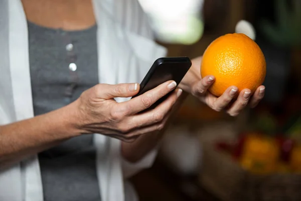 Sección media del cliente femenino sosteniendo naranja mientras usa el teléfono móvil — Foto de Stock