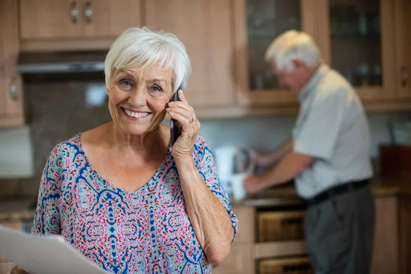 男の台所で作業しながら携帯電話で話している年配の女性 — ストック写真