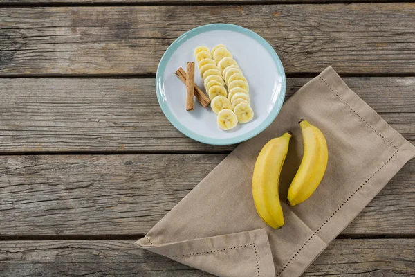 Banana e fette di banana con bastoncino di cannella in piatto su tavolo di legno — Foto Stock