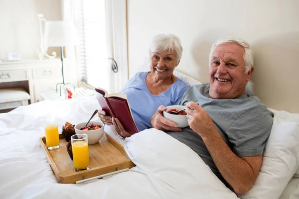 Pareja mayor leyendo un libro mientras desayuna en el dormitorio —  Fotos de Stock