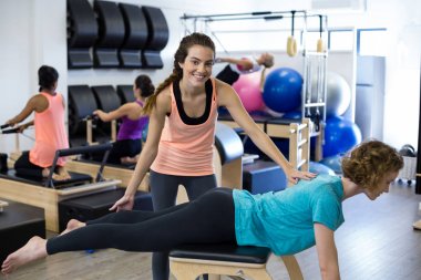 Female trainer assisting woman with stretching exercise on reformer clipart